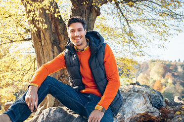 Portrait of smiling young man in a forest sitting on trunk - CHAF001573