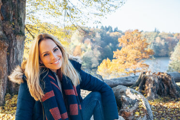 Lächelnde Frau genießt den Herbst in einem Wald auf einem Baumstamm sitzend - CHAF001578