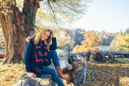 Lächelnde Frau genießt den Herbst in einem Wald auf einem Baumstamm sitzend - CHAF001579
