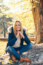 Lächelnde Frau genießt den Herbst in einem Wald auf einem Baumstamm sitzend - CHAF001580