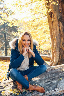 Lächelnde Frau genießt den Herbst in einem Wald auf einem Baumstamm sitzend - CHAF001581