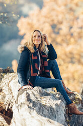 Lächelnde Frau genießt den Herbst in einem Wald auf einem Baumstamm sitzend - CHAF001609