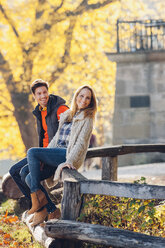 Glückliches Paar sitzt auf einem Holzgeländer in einem herbstlichen Park - CHAF001635