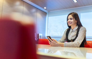 Porträt einer lächelnden Geschäftsfrau, die in einem Konferenzraum mit ihrem Smartphone sitzt - MGOF001346