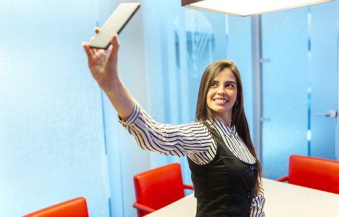 Porträt einer lächelnden Geschäftsfrau, die ein Selfie mit ihrem Smartphone in einem Konferenzraum macht, lizenzfreies Stockfoto