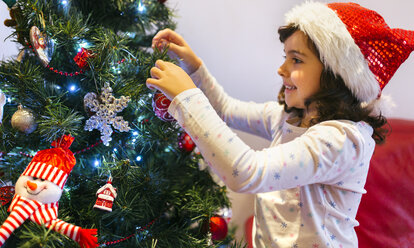 Glückliches kleines Mädchen mit Weihnachtsmütze beim Schmücken des Weihnachtsbaums - MGOF001364