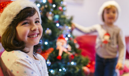 Porträt eines glücklichen Mädchens zur Weihnachtszeit mit ihrer kleinen Schwester im Hintergrund - MGOF001366