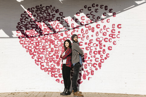 Young couple standing back to back in front of a brick wall with a heart - JRFF000404