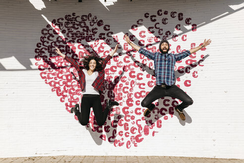 Happy young couple jumping in front of a brick wall with a heart - JRFF000405