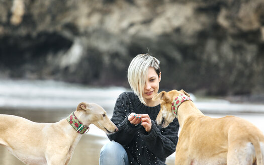 Spanien, Llanes, junge Frau füttert ihre Windhunde am Strand - MGOF001370