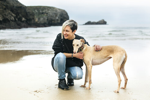 Spanien, Llanes, glückliche junge Frau mit ihrem Windhund am Strand - MGOF001373