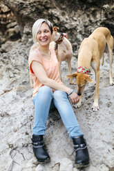 Spain, Llanes, portrait of laughing young woman with her greyhounds on the beach - MGOF001375