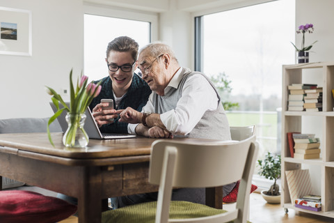 Senior man and his grandson sitting at table in the living room with laptop and smartphone stock photo