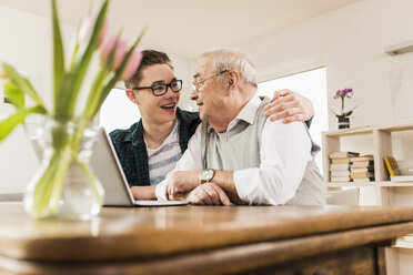 Älterer Mann und sein Enkel sitzen sich am Wohnzimmertisch mit Laptop gegenüber - UUF006566