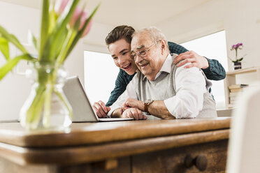 Älterer Mann und sein Enkel schauen gemeinsam auf einen Laptop im Wohnzimmer - UUF006567