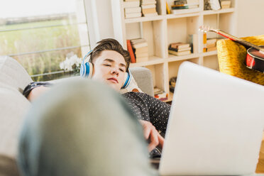 Junger Mann auf der Couch liegend mit Laptop und Kopfhörern - UUF006638