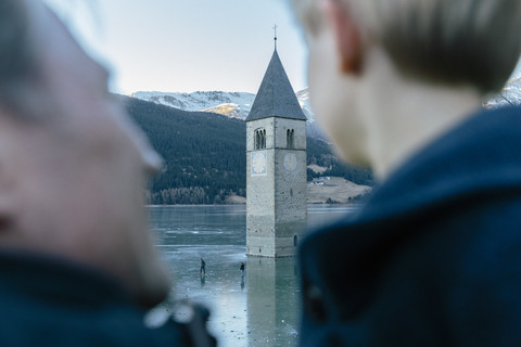 Italien, Vinschgau, Vater und Sohn betrachten die versunkene Turmspitze im zugefrorenen Lago di Resia, lizenzfreies Stockfoto