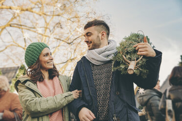 Glückliches Paar mit einem Baum, das über den Weihnachtsmarkt spaziert - MFF002643