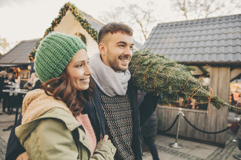 Glückliches Paar mit einem Baum, das über den Weihnachtsmarkt spaziert - MFF002646