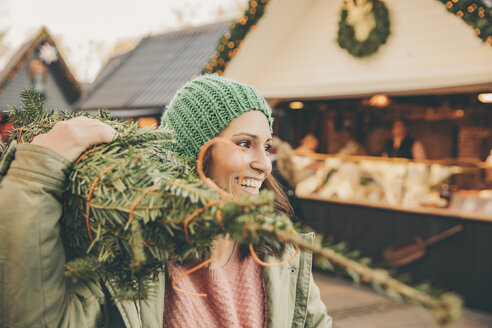 Glückliche Frau mit einem eingepackten Baum, die über den Weihnachtsmarkt läuft - MFF002648