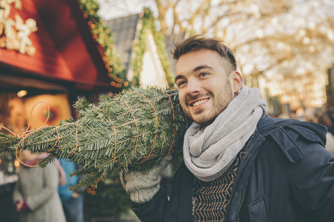 Lächelnder Mann mit einem eingepackten Baum, der über den Weihnachtsmarkt läuft, lizenzfreies Stockfoto