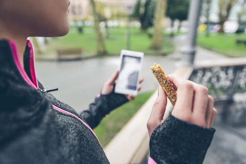 Frau isst Müsliriegel nach dem Training und hält Smartphone, lizenzfreies Stockfoto