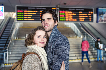 Lächelndes junges Paar, das sich in der Bahnhofshalle umarmt - HAPF000219