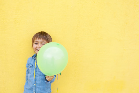 Smiling little boy with green balloon and streamer standing in front of yellow wall stock photo