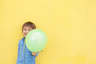 Lächelnder kleiner Junge mit grünem Luftballon und Luftschlange vor gelber Wand stehend - VABF000144