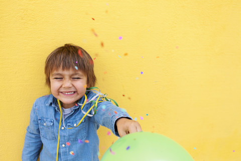 Lächelnder kleiner Junge mit grünem Luftballon, Luftschlange vor gelber Wand stehend, während Konfetti herunterfällt, lizenzfreies Stockfoto