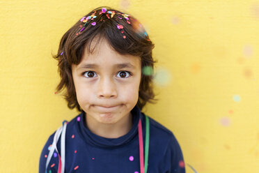 Little boy pulling funny face in front of yellow wall while confetti falling down on him - VABF000146