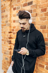 Portrait of young man leaning against brick wall listening music with headphones - MGOF001430