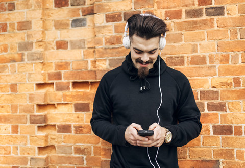 Porträt eines jungen Mannes vor einer Backsteinmauer, der mit Kopfhörern Musik hört, lizenzfreies Stockfoto