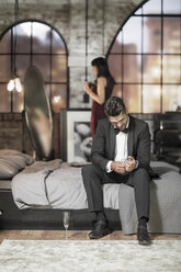 Elegant man on bed getting dressed with wife in background - ZEF008464