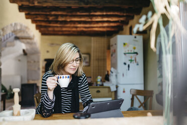 Woman drinking coffee in the kitchen of her farmhouse while using digital tablet - JRFF000431