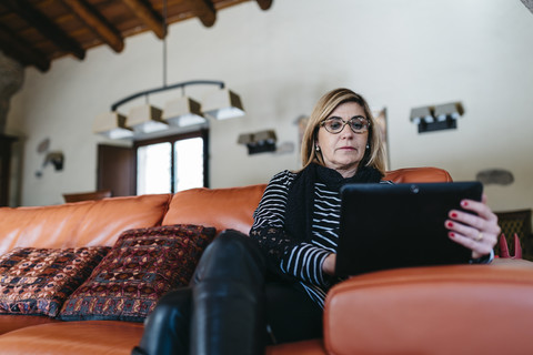 Frau sitzt auf der Couch im Wohnzimmer ihres Bauernhauses und benutzt ein digitales Tablet, lizenzfreies Stockfoto