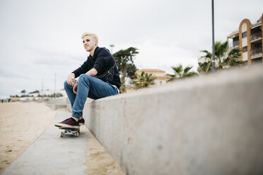Spanien, Torredembarra, lächelnder junger Skateboarder, der auf einer Mauer sitzt - JRFF000434