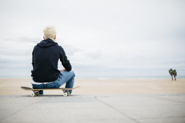 Spanien, Torredembarra, Rücken eines jungen Mannes, der auf seinem Skateboard sitzt und auf das Meer schaut - JRFF000441