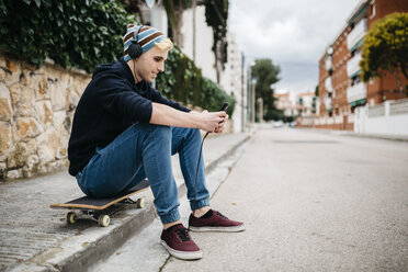 Spanien, Torredembarra, lächelnder junger Mann, der auf seinem Skateboard am Straßenrand sitzt und mit Kopfhörern Musik hört - JRFF000443