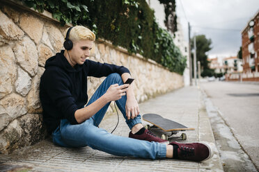 Spanien, Torredembarra, junger Skateboarder sitzt auf dem Bürgersteig und hört mit Kopfhörern Musik - JRFF000444