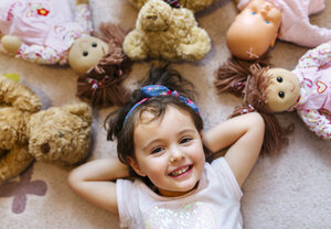 Porträt eines lächelnden kleinen Mädchens, das auf dem Boden liegt und von Teddys und Puppen umgeben ist - MGOF001443