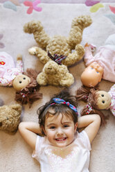 Porträt eines lächelnden kleinen Mädchens, das auf dem Boden liegt und von Teddys und Puppen umgeben ist - MGOF001444