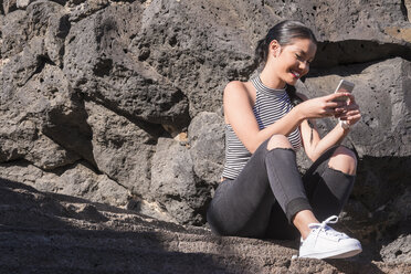 Spain, Tenerife, young woman sitting in front of a rock face looking at her smartphone - SIPF000187