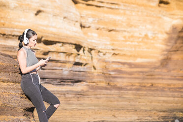 Spain, Tenerife, young woman with headphones standing in front of a rock face looking at her smartphone - SIPF000189