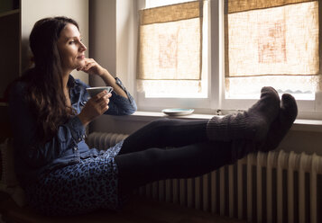 Tagträumende Frau mit Füßen auf der Heizung, die eine Tasse Tee trinkt und durch das Fenster schaut - OPF000108