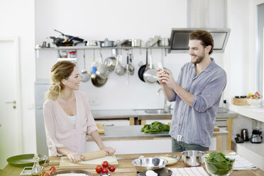 Man taking cell phone picture of smiling woman preparing pizza dough in kitchen - FMKF002370