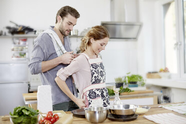 Man helping woman putting on apron in kitchen - FMKF002376