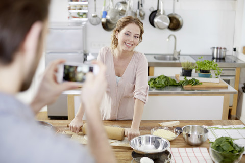 Mann macht Handyfoto von lächelnder Frau, die in der Küche Teig zubereitet, lizenzfreies Stockfoto