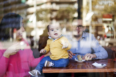 Baby boy sitting on table in a coffee shop looking through windowpane - VABF000199