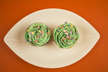 Zwei Cupcakes mit grüner Creme und Backdekor auf einem Teller vor orangefarbenem Hintergrund - ABZF000211